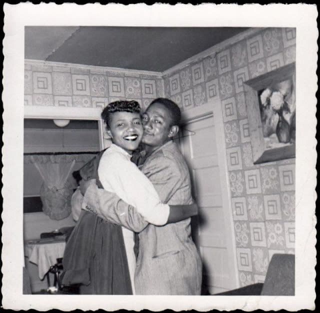Photographie ancienne en noir et blanc d'un couple s'enlaçant en riant, dans un intérieur d'époque.